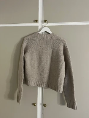 Beige stickad kofta från NA-KD - Mysig beige stickad kofta från NA-KD i storlek L. Koftan har lång ärm, ribbade muddar och v-ringad öppning. Snygga broderade blommor i ljusgult och grönt längs framkanten ger en extra detalj. Perfekt för lager-på-lager under hösten och vintern.