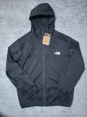 Svart vindjacka från The North Face - Svart vindjacka från The North Face med huva och hel dragkedja framtill. Jackan har en clean look med logga på bröstet och ryggen. Perfekt för blåsiga väder eller för att använda under träning. Hör av dig vid frågar eller funderingar. 