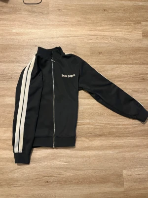 Svart Palm Angels track jacket L - Svart track jacket från Palm Angels med vita ränder längs ärmarna och logga på bröstet. Jackan har hel dragkedja framtill, ribbade muddar och hög krage. Materialet är mestadels polyester med lite bomull i fodret. Perfekt för en sportig streetwear-look.