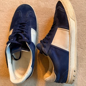 Valentino Flycrew 43 - Snygga Valentino Rockrunner sneakers i mörkblå mocka och vitt skinn. Klassisk låg modell med snörning och platt sula. Sidan av sulan har coola nitar som ger extra edge. Ena skon saknar 3 nitar. Annars jättefint skick. Perfekt för dig som gillar exklusiva och stilrena sneakers med unik detalj.