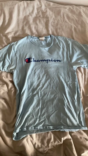 Ljusblå Champion t-shirt - Snygg ljusblå t-shirt från Champion med klassisk logga tryckt framtill i blått och rött. T-shirten har rund halsringning och korta ärmar. Perfekt för en avslappnad och sportig stil.
