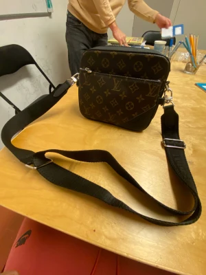 Louis Vuitton axelväska monogram brun - Snygg axelväska från Louis Vuitton i klassiskt monogrammönster. Väskan är brun med svarta detaljer och har justerbar svart axelrem. Tillverkad i skinn med två fack och dragkedjor. Perfekt för dig som vill ha en stilren och ikonisk accessoar. 