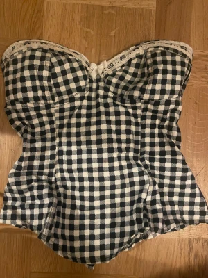 Svartvit rutig korsett med snörning - Säljer en gullig svartvit rutig korsett med snörning i ryggen och en söt rosett framtill. Toppen är justerbar med axelbanden som man antingen kan knyta fast eller ha dom hängande. Och har spetsdetaljer längs överkanten. Materialet känns som bomull och har en klassisk, cool vibe. Perfekt för fest eller bara att ha under en kofta.