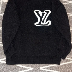 Svart stickad tröja Louis Vuitton - Svart stickad tröja från Louis Vuitton med stor vit LV-logga på bröstet. Tröjan har rund hals, ribbade muddar och lång ärm. Materialet är mjukt och värmande, perfekt för kyliga dagar. Snygg och stilren design som sticker ut med den ikoniska loggan.