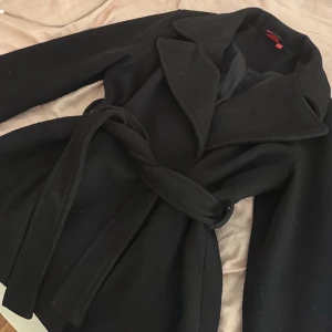 Svart kappa från RYVLS med bälte - Snygg svart kappa från RYVLS med bred krage och knytbälte i midjan. Jackan har en klassisk och stilren look, perfekt för dig som gillar en clean och elegant stil. Materialet känns mjukt och värmande, och modellen är normal i passformen.