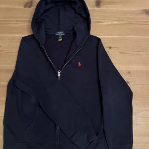 Svart hoodie från Polo Ralph Lauren - Svart hoodie från Polo Ralph Lauren med dragkedja och huva. Klassisk röd logga broderad på bröstet. Tröjan har långa ärmar och två fickor framtill. Mjuk insida i bomullsmix som gör den extra skön att bära. (Använd ungefär tre gånger) Hör av er vid minsta lilla fråga mvh Victor!