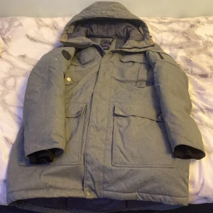 Grå vinterjacka från Everest, strl 38 - Säljer en grå vinterjacka från Everest i storlek 38. Jackan har huva, flera stora fickor med lock och snyggt Everest-märke på ärmen. Perfekt för kalla dagar med sin långa modell och rejäla material. Stängs med dragkedja och knappar framtill.