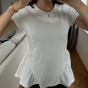 Vit blus med volang nertill - Vit blus med rund hals och korta ärmar från Gina tricot. Toppen har en snygg volangdetalj nertill som ger en feminin och trendig look. Materialet känns mjukt och bekvämt, perfekt för dig som gillar stilrena och enkla plagg med en twist.