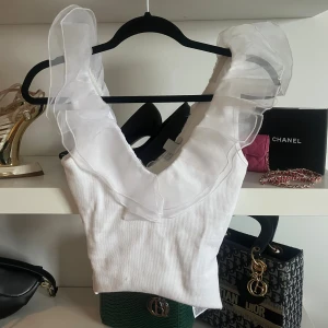 Vit ärmlös topp med volang - Säljer en vit ärmlös topp med bred v-ringning och transparent volangdetalj runt halsen. Toppen har en stilren och feminin look med ribbad struktur i tyget och volangen ger en extra touch. Perfekt för dig som gillar att sticka ut med detaljer. Är en body från början.