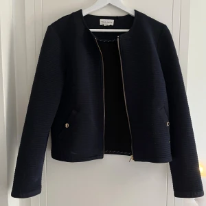 Mörkblå jacka  - Stilren mörkblå  tunnare  från Holly & White med diskret ribbad struktur och guldfärgade detaljer vid fickorna samt dragkedjan. Jackan har rund halsringning, dragkedja framtill och långa ärmar. Perfekt för enklare uppklädd outfit. Säljer då den inte längre används. 