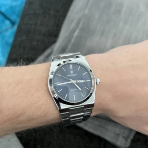 Herrklocka  - Stilren med blå urtavla och dag/datumvisning. Boetten och länken är i blankt silverfärgat metall, vilket ger en modern och clean look. Klockan har quartzverk och minimalistiska indexmarkeringar. Perfekt accessoar för dig som gillar klassisk design.