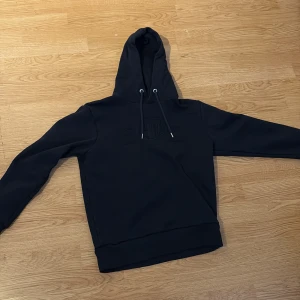 Mörk blå hoodie från GANT med broderi - Mörk blå hoodie från GANT med broderad logga och sköld på bröstet. Klassisk modell med huva och snörning, ribbade muddar och mjukt material. Perfekt för dig som gillar stilren och sportig look.