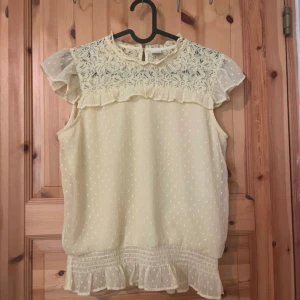 Söt ljusgul blus med spets och volang - Ljusgul blus med spets upptill, volangdetaljer vid axlar och nederkant samt diskret prickigt mönster. Ärmlös modell med romantisk känsla och knäppning bak i nacken. Perfekt för vår och sommar.