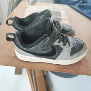 Grå Nike sneakers med kardborre - Snygga grå och svarta Nike sneakers med klassisk swoosh på sidan. Skorna har både snörning och praktisk kardborre för enkel påtagning. Ovandelen är i syntet och skinn, med perforerad tå för extra ventilation. Perfekt för dig som gillar sportig stil.