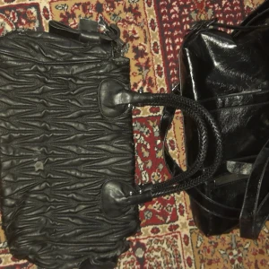 Miu Miu svart handväska i skinn - Cool svart handväska från Miu Miu i skinn med snyggt rynkad struktur och flätade handtag. Väskan har metallogo framtill och detaljer i silver. Perfekt för dig som vill ha en unik och trendig accessoar med plats för allt du behöver.