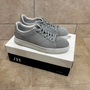 Gråa selected homme skor - Snygga grå sneakers från Selected Homme i mjuk mocka med klassisk rund tå och platt vit sula. Skorna har matchande grå snörning och en stilren, minimalistisk design som passar till det mesta. Perfekta för dig som gillar en clean och modern look. Jag har använt dom2 gånger men dom ser helt sprit nya ut.