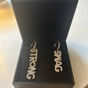 Glittriga örhängen med text SWAG & STRONG - Säljer ett par coola örhängen i silverfärg med glittriga stenar. Ena örhänget har texten 'SWAG' och det andra 'STRONG' i stora bokstäver. Perfekt för dig som vill sticka ut och visa din personlighet. 