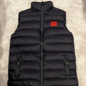 Svart dunväst från Hugo Boss - Snygg svart dunväst från Hugo Boss med röd HUGO-logga på bröstet och vit HUGO-text på kragen. Västen har quiltad design, två fickor med dragkedja och hög krage. Perfekt för att addera lite edge till din outfit.