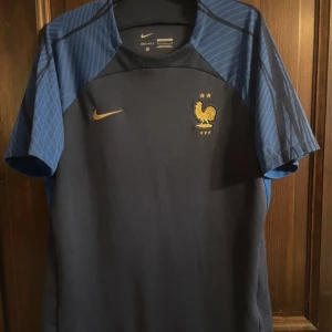Frankrike fotbollströja Nike blå L - Säljer en officiell Frankrike fotbollströja från Nike i storlek L. Tröjan är mörkblå med ljusblå ärmar och har guldiga detaljer som Nike-logga och landslagets emblem med tupp och två stjärnor. Tillverkad i lätt och ventilerande material, perfekt för match eller träning.