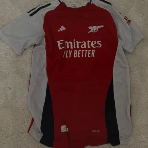 Arsenal Ødegaard 8 matchtröja Adidas - Arsenal hemmatröja från Adidas med Ødegaard 8 på ryggen. Klassisk röd front med vita ärmar och svarta detaljer. Broderad klubbmärke och sponsortryck på bröstet. Tillverkad i lätt och ventilerande polyester, perfekt för match eller träning.