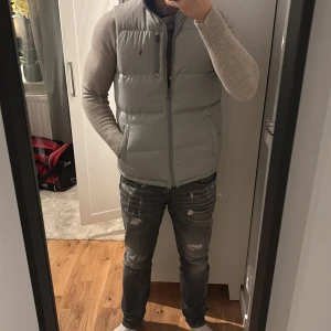 Vändbar dunväst från Polo Ralph Lauren - Snygg vändbar dunväst från Polo Ralph Lauren med en sida i ljusgrått och en i klarblått. Västen har ståkrage, två sidofickor och en bröstficka med dragkedja på den gråa sidan. Ikoniska Polo-loggan broderad på båda sidor. Perfekt för lager-på-lager och streetstyle.