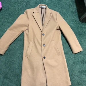 Beige kappa från ZARA - Snygg beige kappa från ZARA med klassisk krage och tre svarta knappar framtill. Jackan har två fickor på sidorna och är helfodrad. Perfekt för dig som gillar stilrena och tidlösa plagg med enkel design.