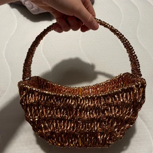 Glittrig handväska i kopparfärgad syntet - Unik handväska med glittrig kopparfärgad yta, flätad i syntetmaterial. Väskan har en rundad form och ett rejält handtag, perfekt för att sticka ut. Botten är förstärkt med rosa papper och gula detaljer. Väskan är riktigt cool och har en trendig vibe.