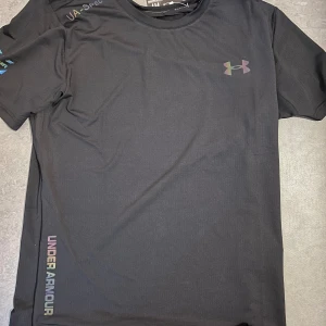 Svarta träningsbyxor från Under Armour - Snygga svarta träningsbyxor från Under Armour med elastisk midja och diskret logga i regnbågsskimrande färg på höften. Byxorna har en sportig passform och är gjorda i lätt och stretchigt syntetmaterial som andas bra under träning.