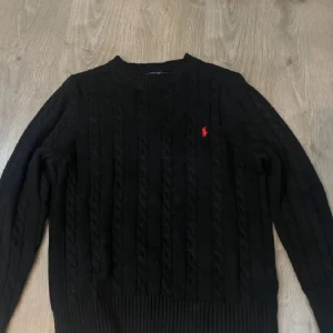 Svart stickad tröja från Ralph Lauren - Svart kabelstickad tröja från Ralph Lauren med klassisk röd logga broderad på bröstet. Rund hals och ribbade muddar vid ärmslut och nederkant. Perfekt för dig som gillar stilrena och tidlösa plagg.