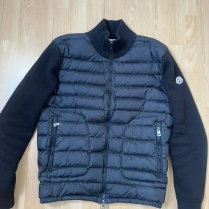 Svart Cardigan från Moncler - Säljer en svart pufferjacka från Moncler med quiltad framsida och stickade ärmar. Jackan har två sidofickor med dragkedja, hög krage och Moncler-logga på ärmen. Materialet är en mix av dun och stickad textil, perfekt för kyliga dagar.