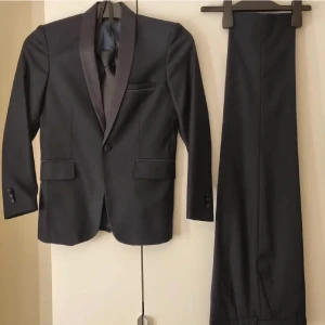 Mörkblå kostym med väst och slips för barn - Säljer en stilren mörkblå kostym med matchande väst och slips. Kavajen har smala slag i satin, två fickor med lock och en bröstficka. Byxorna är raka och klassiska. Västen har tre knappar och två fickor. Perfekt för dig som vill ha en elegant och tidlös look. Storlek 134/140