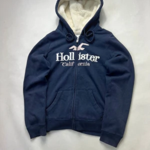 Marinblå hoodie från Hollister - Säljer en marinblå hoodie från Hollister med vit fluffig huva och broderad logga på bröstet. Tröjan har dragkedja, snörning vid huvan och två fickor framtill. Perfekt för dig som gillar en chill och snygg stil.