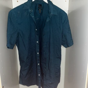 Mörkblå kortärmad skjorta H&M - Kortärmad skjorta från H&M i mörkblå/grön färg med små vita prickar över hela tyget. Slim fit-modell med klassisk krage och knappar framtill. Perfekt för dig som gillar en stilren men ändå lekfull look.