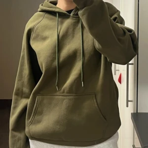 Mörkgrön hoodie med huva och ficka - Säljer en mörkgrön/olivgrön hoodie med huva och snörning. Tröjan har en stor magficka framtill och långa ärmar med muddar. Storlek M, passar på en S om man vill ha oversize.