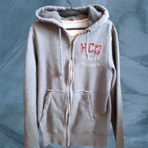 Grå hoodie från Hollister med dragkedja - Säljer en grå hoodie från Hollister med broderad HCO-text på bröstet. Tröjan har huva med snörning, två stora fickor framtill och dragkedja hela vägen. Perfekt för chill dagar och snygg streetstil.