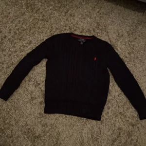 Mörkblå stickad tröja Polo Ralph Lauren - Mörkblå stickad tröja från Polo Ralph Lauren med klassisk kabelstickning och röd broderad logga på bröstet. Tröjan har rund halsringning, ribbade muddar och lång ärm. Perfekt för dig som gillar stilren och tidlös design.