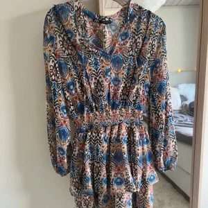 Mönstrad kortklänning från ZARA - Säljer en färgstark kortklänning från ZARA med blommigt och geometriskt mönster i blått, rött, beige och svart. Klänningen har långärmade ärmar, v-ringning och smock i midjan för en snygg passform. Perfekt för dig som gillar boho-vibe och unika prints.