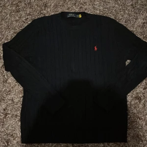 Svart stickad tröja från Polo Ralph Lauren - Svart stickad tröja från Polo Ralph Lauren med klassisk röd logga broderad på bröstet. Tröjan har rund halsringning, diskret ribbad struktur och långa ärmar. Perfekt för dig som gillar stilrena och tidlösa plagg.