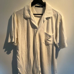Vit frottéskjorta från Zara Man - Vit skjorta från Zara Man i mjuk frotté med klassisk krage, korta ärmar och bröstficka. Skjortan har knappar framtill och en relaxed passform som ger en chill vibe. Perfekt för dig som gillar stilren och enkel design.