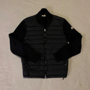 Mörkblå väst  - Mörkblå långärmad väst med stickade ärmar och quiltad kropp. Jackan har dragkedja framtill och Moncler-logga på vänster ärm. Perfekt för dig som vill ha en stilren och varm jacka med snygga detaljer.