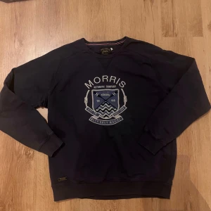Mörkblå sweatshirt från Morris XL - Snygg mörkblå sweatshirt från Morris i storlek XL med broderat marin-inspirerat emblem och text på bröstet. Tröjan har rund hals, ribbade muddar och är tillverkad i mjuk bomull. Perfekt för dig som gillar klassisk och sportig stil.