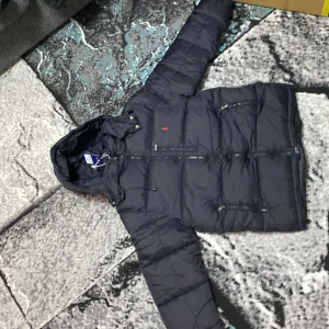 Svart pufferjacka från Polo Ralph Lauren - Svart pufferjacka från Polo Ralph Lauren med huva och dragkedja framtill. Jackan har flera praktiska fickor med dragkedja och den klassiska röda Polo-loggan broderad på bröstet. Perfekt för kalla dagar och har en snygg, sportig look.