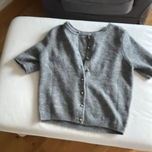 Grå stickad kofta från H&M - Snygg grå stickad kofta från H&M med korta ärmar och rund hals. Koftan har guldfärgade knappar framtill och ribbade muddar vid hals, ärmslut och nederkant. Perfekt att slänga över en t-shirt eller linne för en chill look.