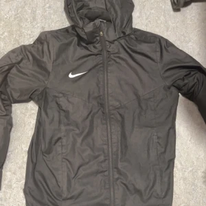 Svart vindjacka från Nike med huva - Svart vindjacka från Nike med vit logga på bröstet. Jackan har huva, hel dragkedja framtill och långärmade ärmar. Materialet är lätt och syntetiskt, perfekt för blåsiga dagar. Enkel och stilren design som funkar till streetwear.