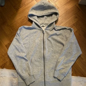 Grå zip hoodie  - Säljer en enkel och clean grå hoodie från C i storlek 146/152. Tröjan har huva, hel dragkedja framtill och två fickor. Materialet är mjukt och skönt, perfekt för chill dagar. Passformen är normal och ärmarna är långa. Snygg att matcha med jeans eller joggers.