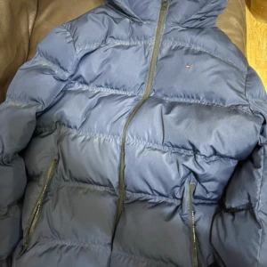 Blå pufferjacka från Tommy Hilfiger - Säljer en blå pufferjacka från Tommy Hilfiger med huva och dragkedja framtill. Jackan har två sidofickor med dragkedja och diskret logga på bröstet. Perfekt för kalla dagar och har en snygg, modern look. Storlek 176.