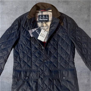 Barbour Sander - Quiltad mörkblå jacka från Barbour med klassisk krage i brun mocka, rutigt foder och snygga detaljer. Jackan har stora fickor med knappar, dragkedja och knappstängning framtill. Perfekt för dig som gillar stilren och tidlös design.