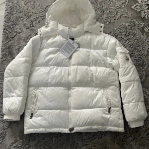 Vit pufferjacka från Moncler, S - Snygg vit pufferjacka från Moncler i storlek S. Jackan har huva, dragkedja framtill och två fickor med dragkedja. Klassisk Moncler-logga på ärmen och coolt serietidningsfoder på insidan. Perfekt för kalla dagar och riktigt stilren look.