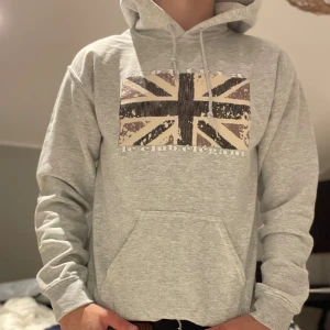 Grå hoodie från Montéva  - Snygg grå hoodie från Le Club Élégant med tryck av en vintage-inspirerad brittisk flagga på bröstet. Tröjan har huva med snörning och en stor magficka. Perfekt för dig som gillar streetstyle och vill sticka ut med coolt tryck.