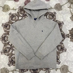Grå Polo Ralph Lauren hoodie - Superfin grå hoodie från Polo Ralph Lauren i mjukt och bekvämt bomullsmaterial. Klassisk modell med huva, känguruficka och den ikoniska marinblå loggan på bröstet. Storlek XL (18–20), men motsvarar ungefär en vanlig S. Använd ett fåtal gånger och i mycket bra skick.  Längd från axeln och ner: 61 cm Bredd under armhålorna: 42 cm  📦 Skickas snabbt & välpackat.
